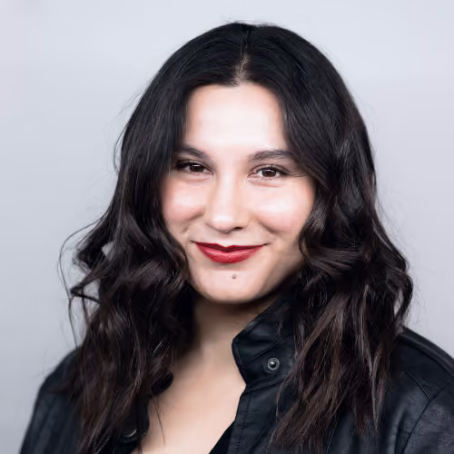 Smiling woman with long wavy dark hair wearing a black leather jacket against a light gray background.