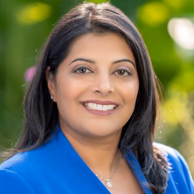 Smiling woman with dark hair wearing a blue blazer and necklace against a green blurred background.