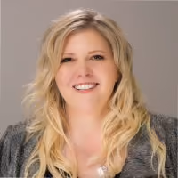Smiling woman with long blonde wavy hair wearing a dark patterned top against a neutral background.