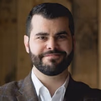Man with dark hair and beard wearing a brown checked blazer and white shirt smiling at the camera.
