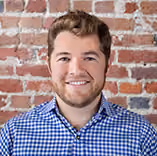 Smiling man with short brown hair and beard wearing a blue checkered shirt against a brick wall.