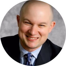 Smiling bald man wearing a suit and blue patterned tie against a gray background.