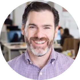 Smiling man with dark hair and beard wearing a purple checkered shirt in an office.