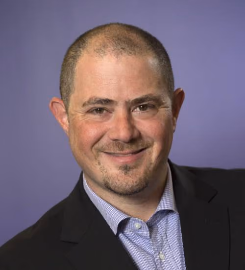 Smiling man with short hair and goatee wearing a black jacket and checked shirt against purple background.