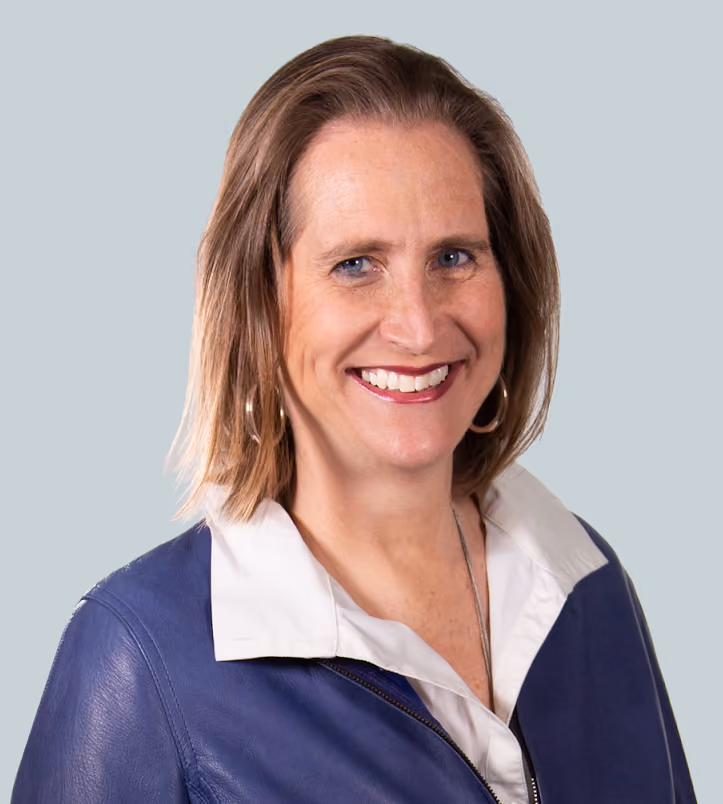 Smiling woman with light brown hair wearing a blue jacket and white collared shirt on gray background.