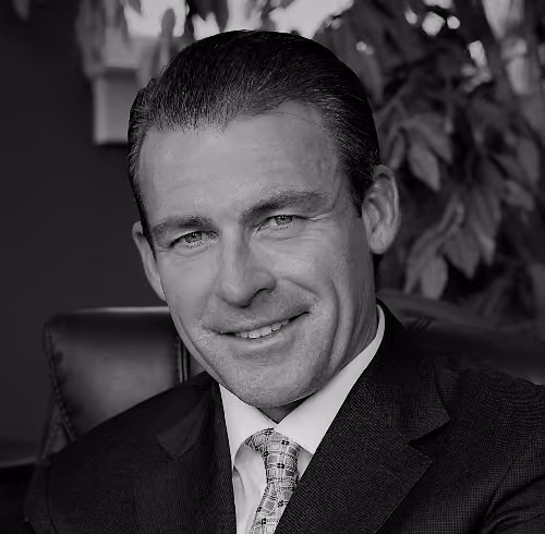 Smiling man in a suit and tie seated in a leather chair with plants in the background.