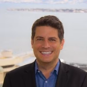 Smiling man in a black blazer and blue shirt standing outdoors with a blurred waterfront background.