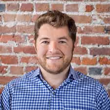 Smiling man with short brown hair and beard wearing a blue checkered shirt in front of a brick wall.