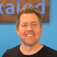 Smiling man with short hair and beard wearing a black shirt in front of a blue wall and wooden sign.