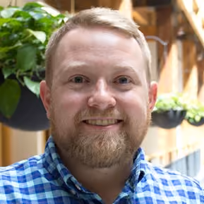 Smiling man with blonde hair and beard wearing a blue checkered shirt indoors with plants in the background.