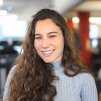 Smiling woman with long curly hair wearing a gray sweater in an office setting.