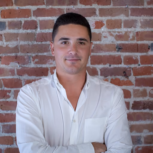 Man with short dark hair wearing a white buttoned shirt stands against a brick wall, smiling