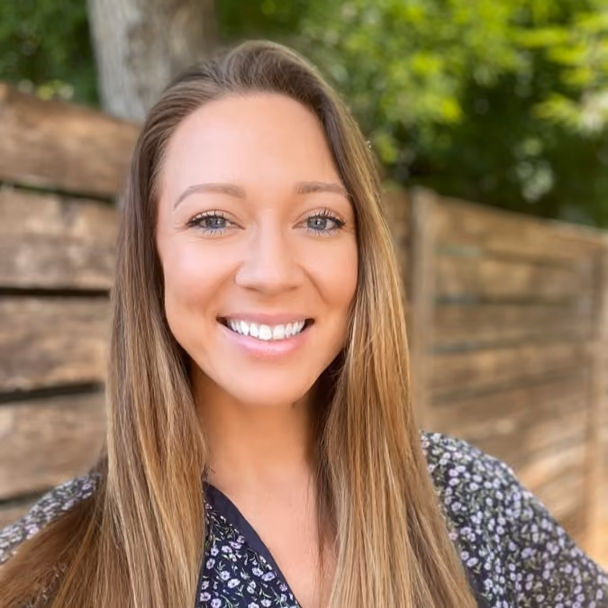 Smiling woman with long light brown hair wearing a floral top standing outdoors by wooden fence.