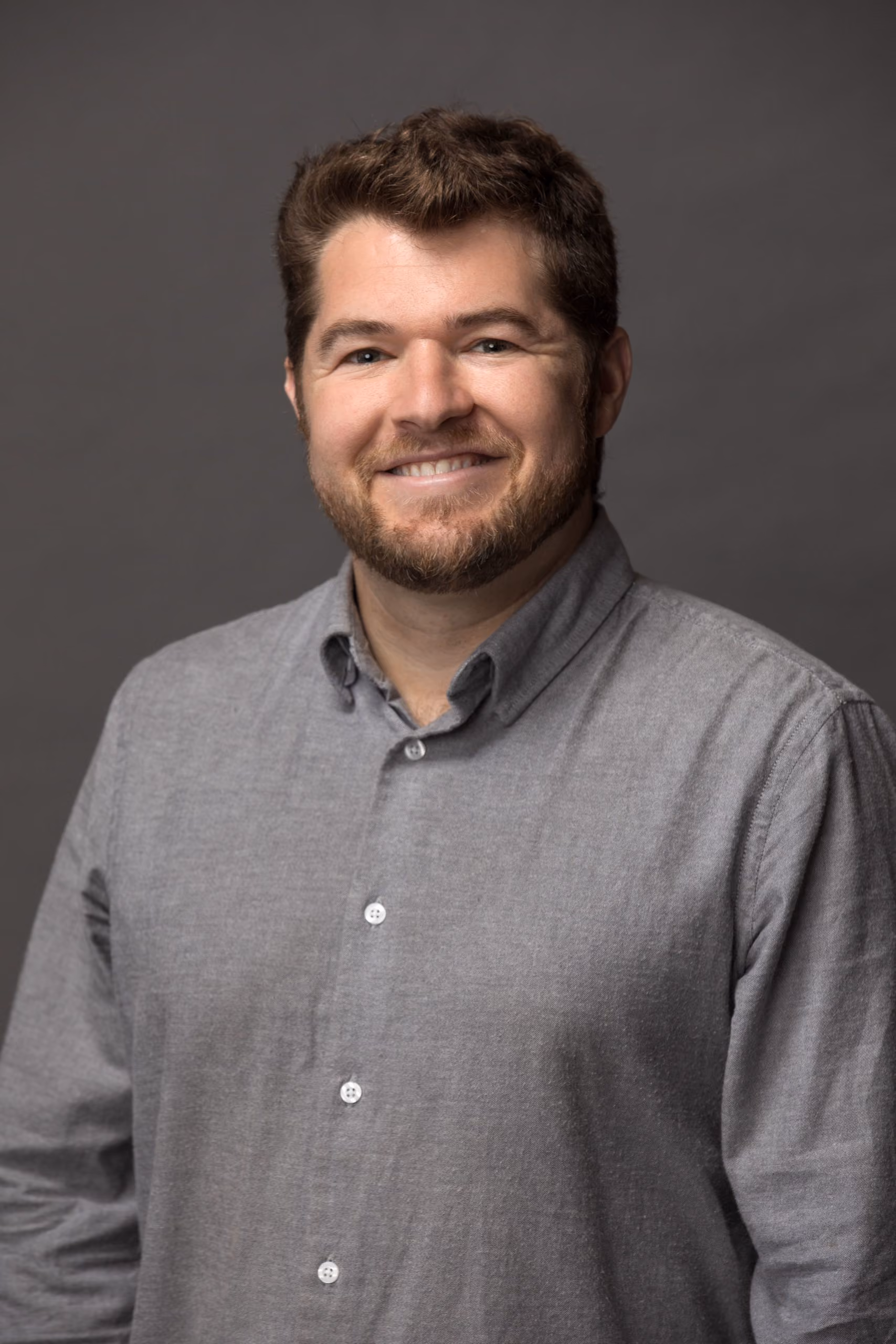 Smiling man with short brown hair and beard wearing a gray button-up shirt against a gray background.