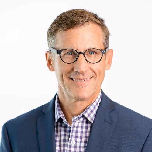 Smiling man with glasses wearing a blue blazer and purple checkered shirt on white background.