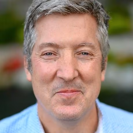 Close-up of a middle-aged man with short gray hair smiling slightly outdoors in soft focus.