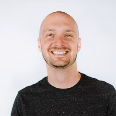 Smiling bald man with a trimmed beard wearing a black shirt against a plain white background.