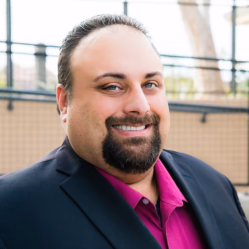 Smiling man with a dark goatee wearing a magenta shirt and black jacket outdoors.