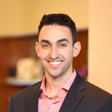 Smiling man with short dark hair wearing a black suit jacket and pink shirt in an indoor setting.