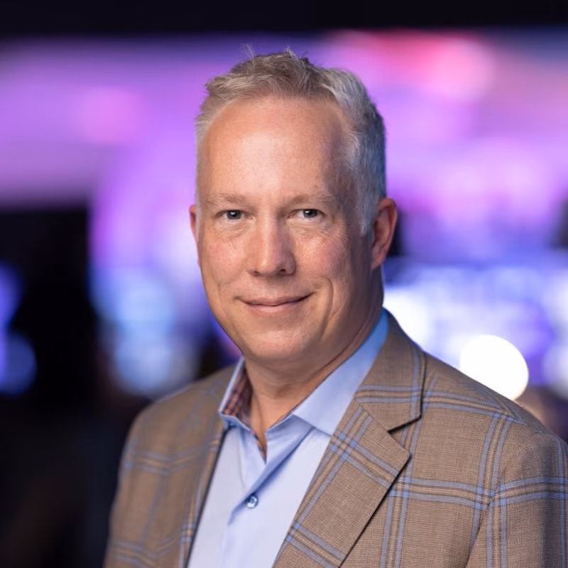 Middle-aged man with gray hair wearing a plaid blazer and light blue shirt with a blurred colorful background.