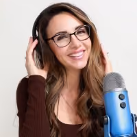 Smiling woman with glasses wearing headphones and speaking into a blue microphone against white background.