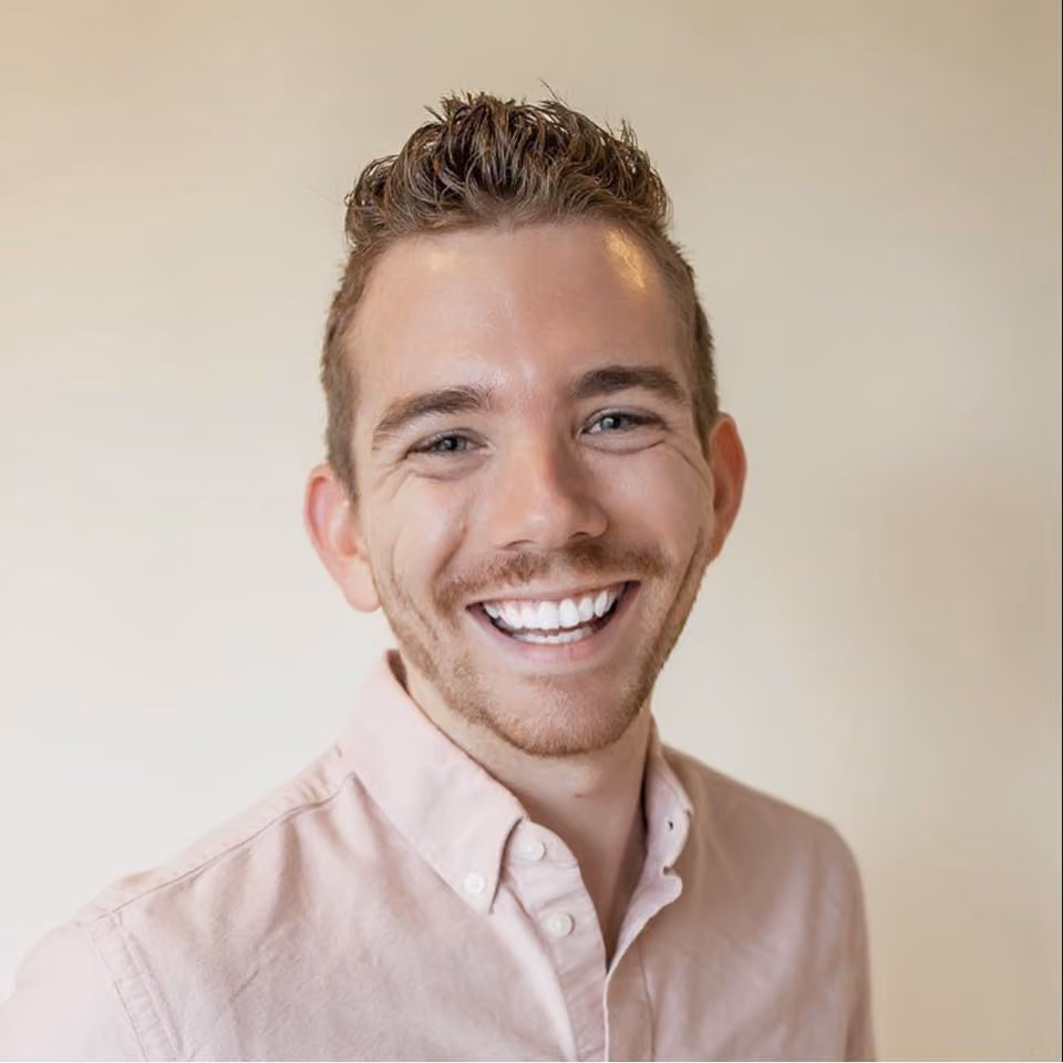 Smiling young man with short curly hair wearing a light pink button-up shirt against a plain background.