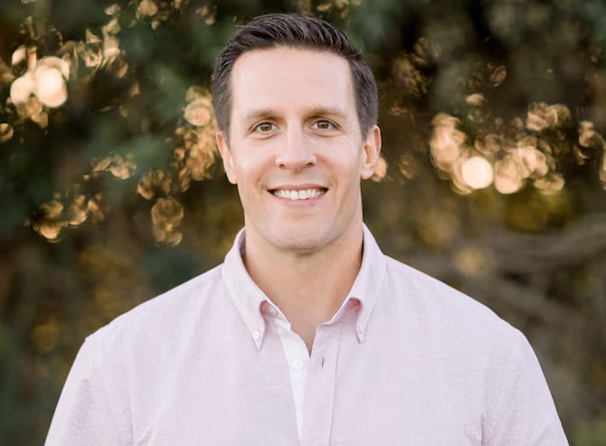 Smiling man with short brown hair wearing a light pink button-up shirt, outdoors with greenery background.