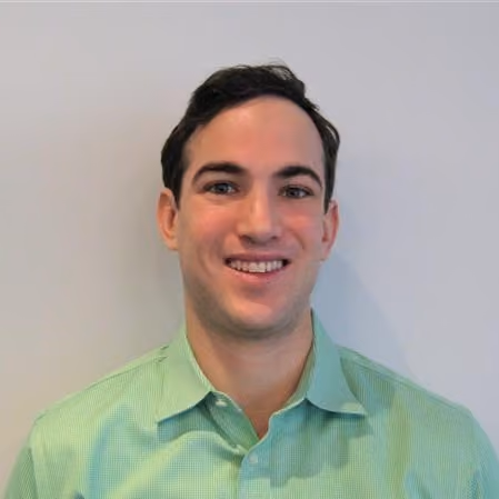 Smiling man with short dark hair wearing a light green collared shirt against a plain wall.