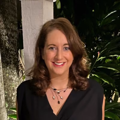 Smiling woman with curly brown hair wearing a black top and necklace, standing outdoors near trees.