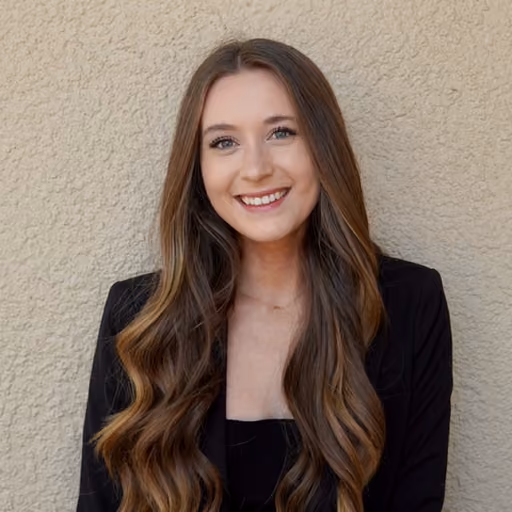 Smiling young woman with long wavy brown hair wearing a black blazer against a textured beige wall.