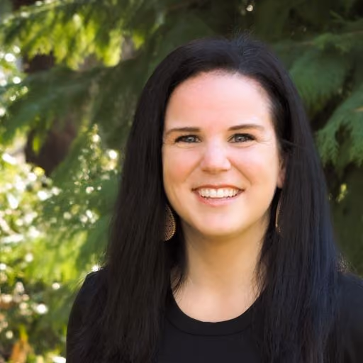 Smiling woman with long black hair standing outdoors with green foliage in the background.
