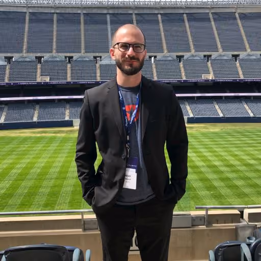 Man with glasses and beard wearing a black suit stands in front of an empty stadium field.