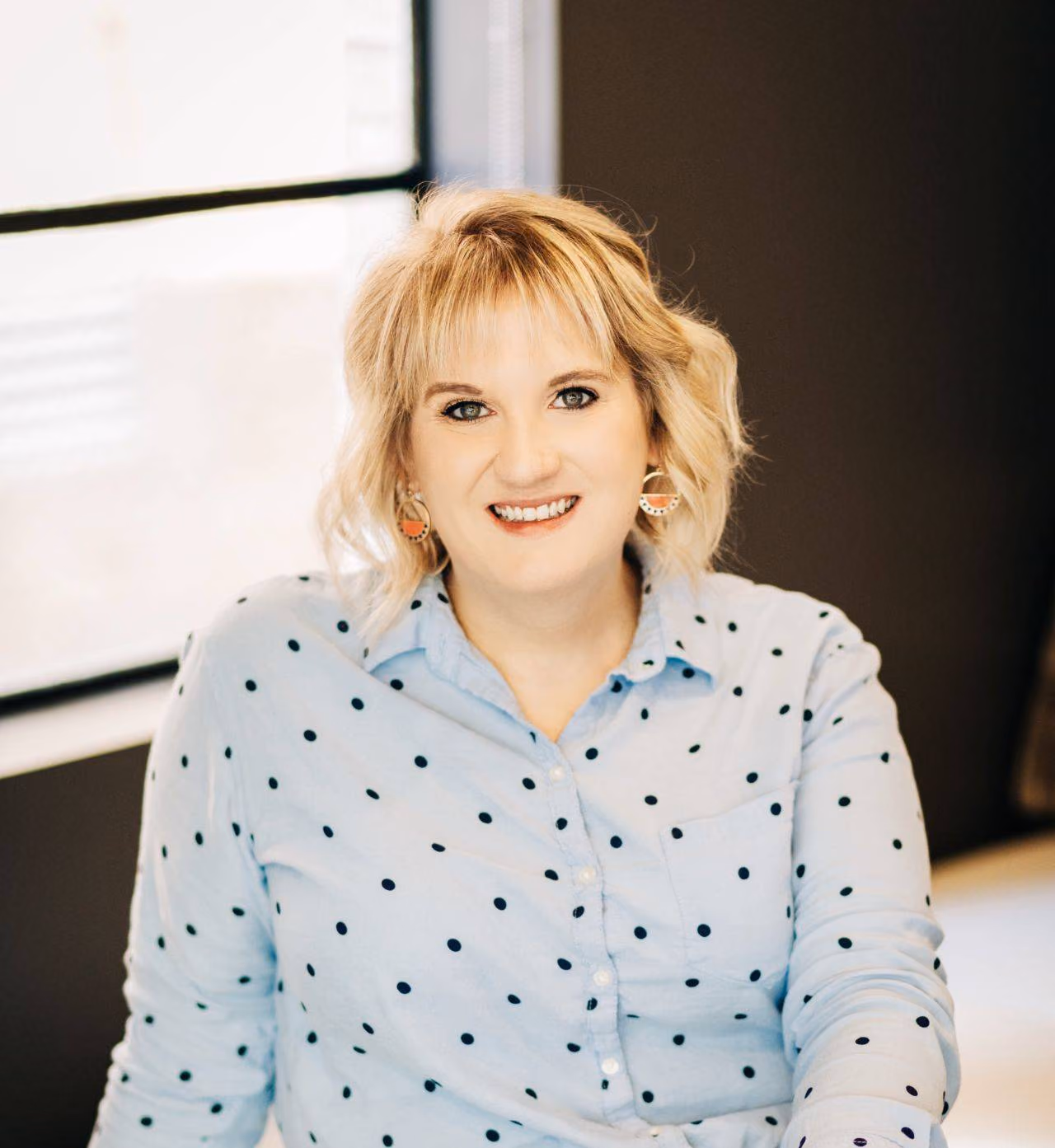 Smiling woman with short blonde hair wearing a blue shirt with black polka dots by a window.