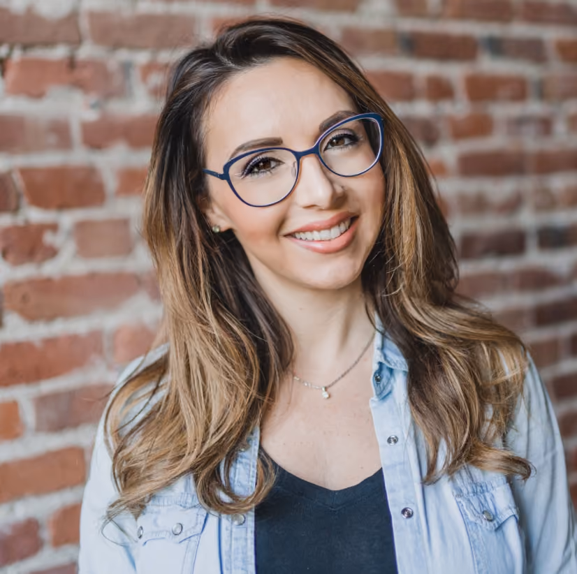 Smiling woman with long hair and blue glasses wearing a denim jacket against a brick wall.