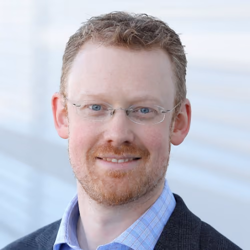 Headshot of a smiling man with light skin, glasses, curly hair, and a short beard wearing a checked shirt and blazer.