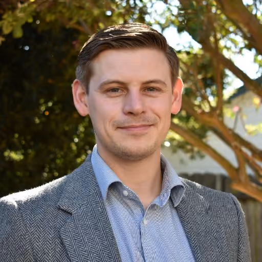 Smiling man in a herringbone jacket and striped shirt outdoors with sunlit trees in the background.