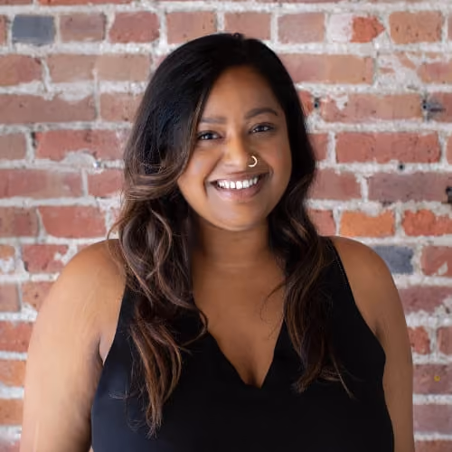 Smiling woman with long dark hair, nose ring, and black sleeveless top against a brick wall.