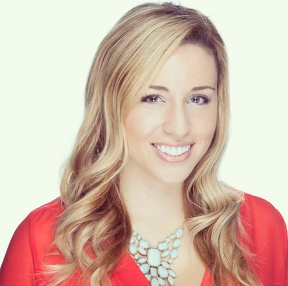 Smiling blonde woman with wavy hair wearing a red blouse and a statement necklace.