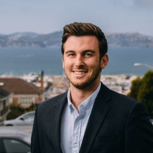 Smiling man in a blazer and light blue shirt with a city and water backdrop.