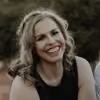 Smiling woman with curly hair wearing a sleeveless black top outdoors with blurred greenery background.