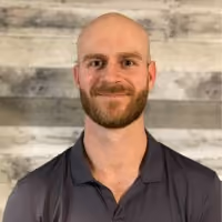 Smiling bald man with a beard wearing a dark gray polo shirt against a wooden background.