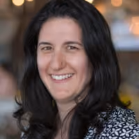 Smiling woman with long dark hair wearing a patterned top with blurred background lights.