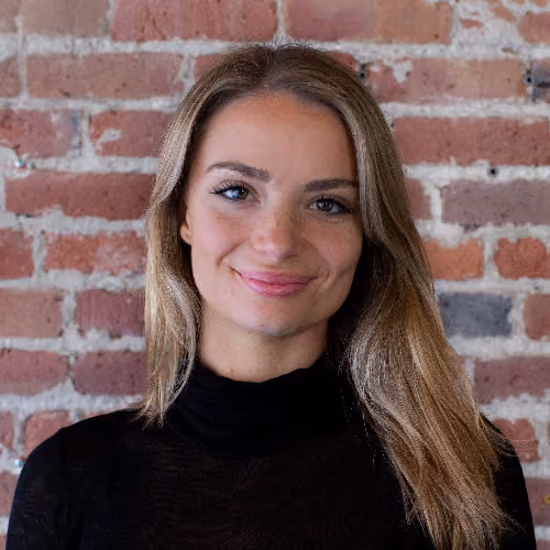 Smiling woman with long blonde hair wearing black top against a brick wall background.