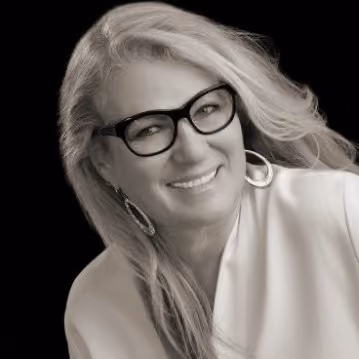 Smiling woman with long hair wearing glasses, hoop earrings, and a light-colored top on a black background.