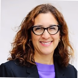 Woman with curly brown hair wearing black glasses, a black blazer, and a purple top smiling.