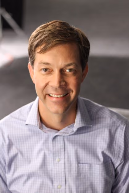 Smiling man with short brown hair wearing a light checkered shirt against a blurred background.