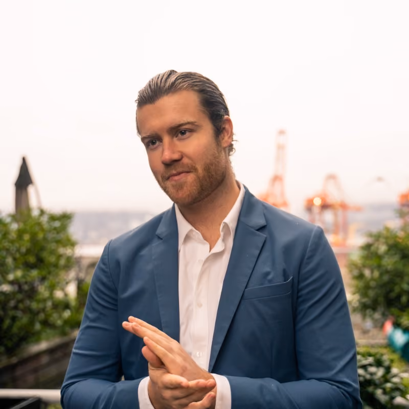 Man with slicked-back hair and beard wearing a blue blazer over a white shirt outdoors.