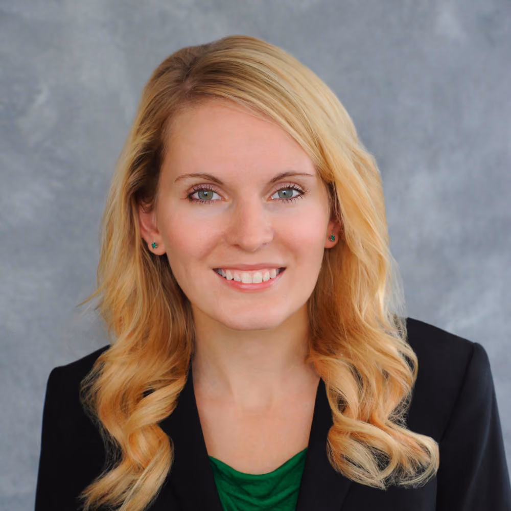 Smiling woman with long blonde hair wearing a black blazer and green top against a gray backdrop.