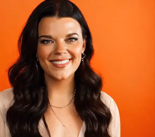 Woman with long wavy dark hair and hoop earrings smiling against an orange background.
