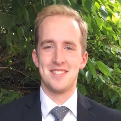 Smiling young man in a suit and polka dot tie with leafy green background.